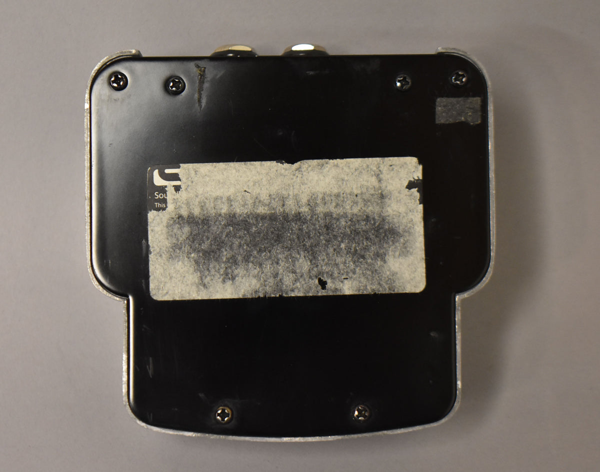 The pedal's black metal baseplate is visible, with a large, worn white sticker covering most of its surface and four visible screws.