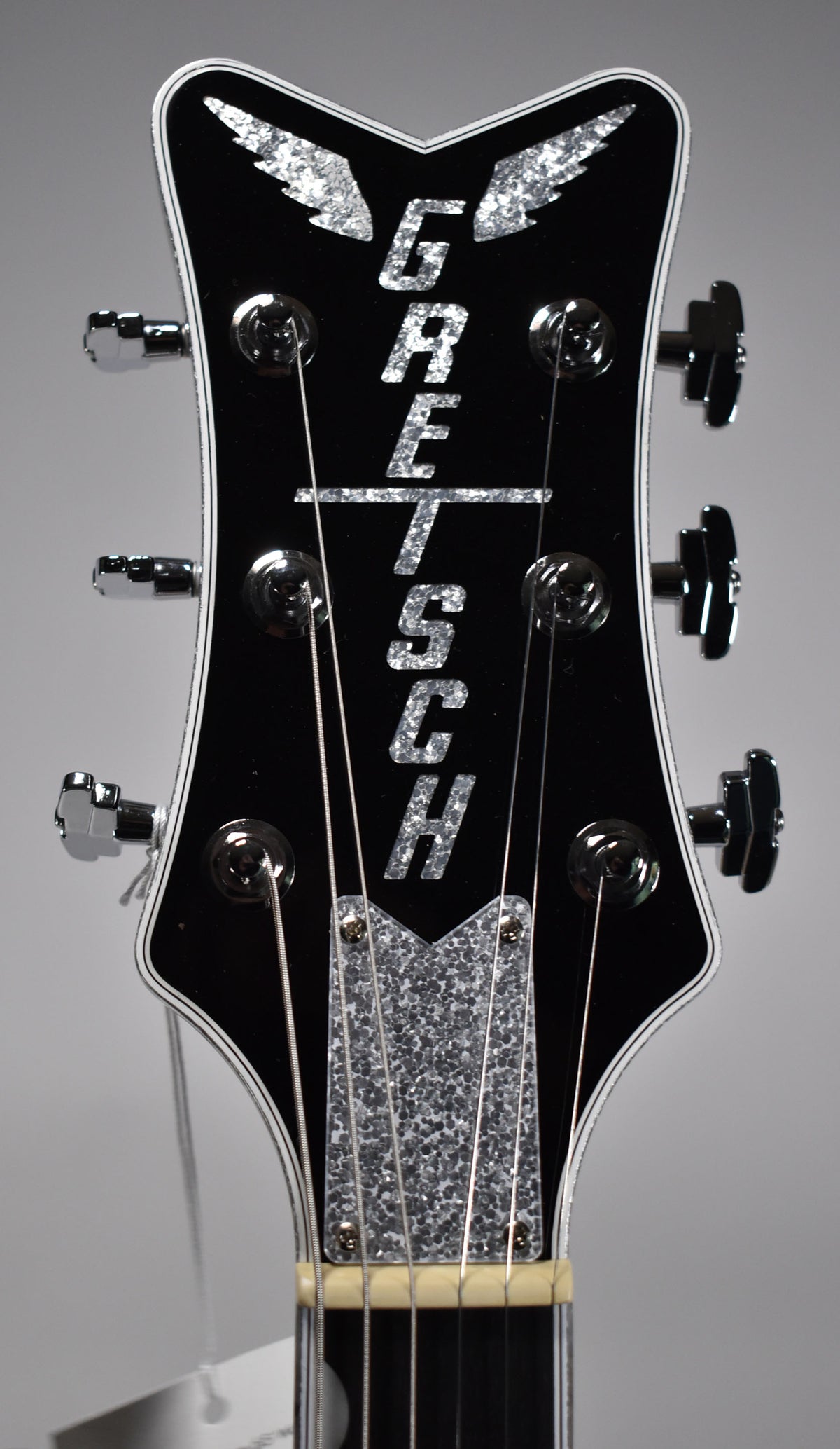 Close-up of the black headstock with white binding, showing six chrome tuners, a silver sparkle truss rod cover, and a bone nut.