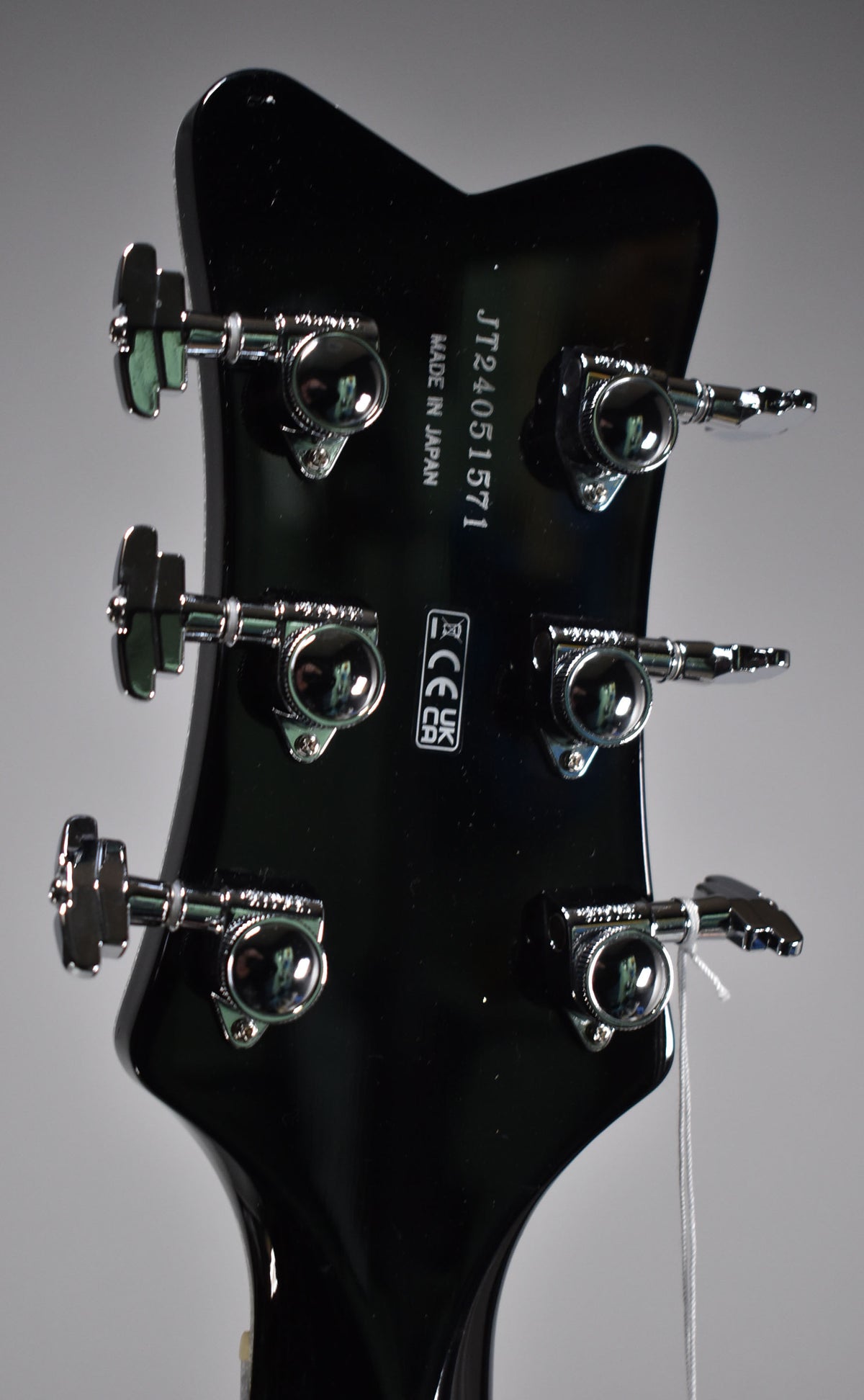 The back of a glossy black headstock with six chrome tuning machines, three on each side, and a white hang tag string.