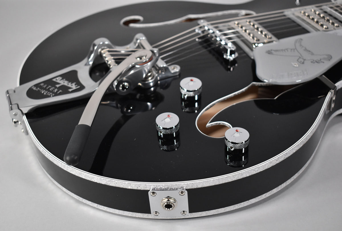 Close-up of a glossy black hollow-body guitar with a Bigsby tremolo, four chrome knobs, and a silver pickguard.