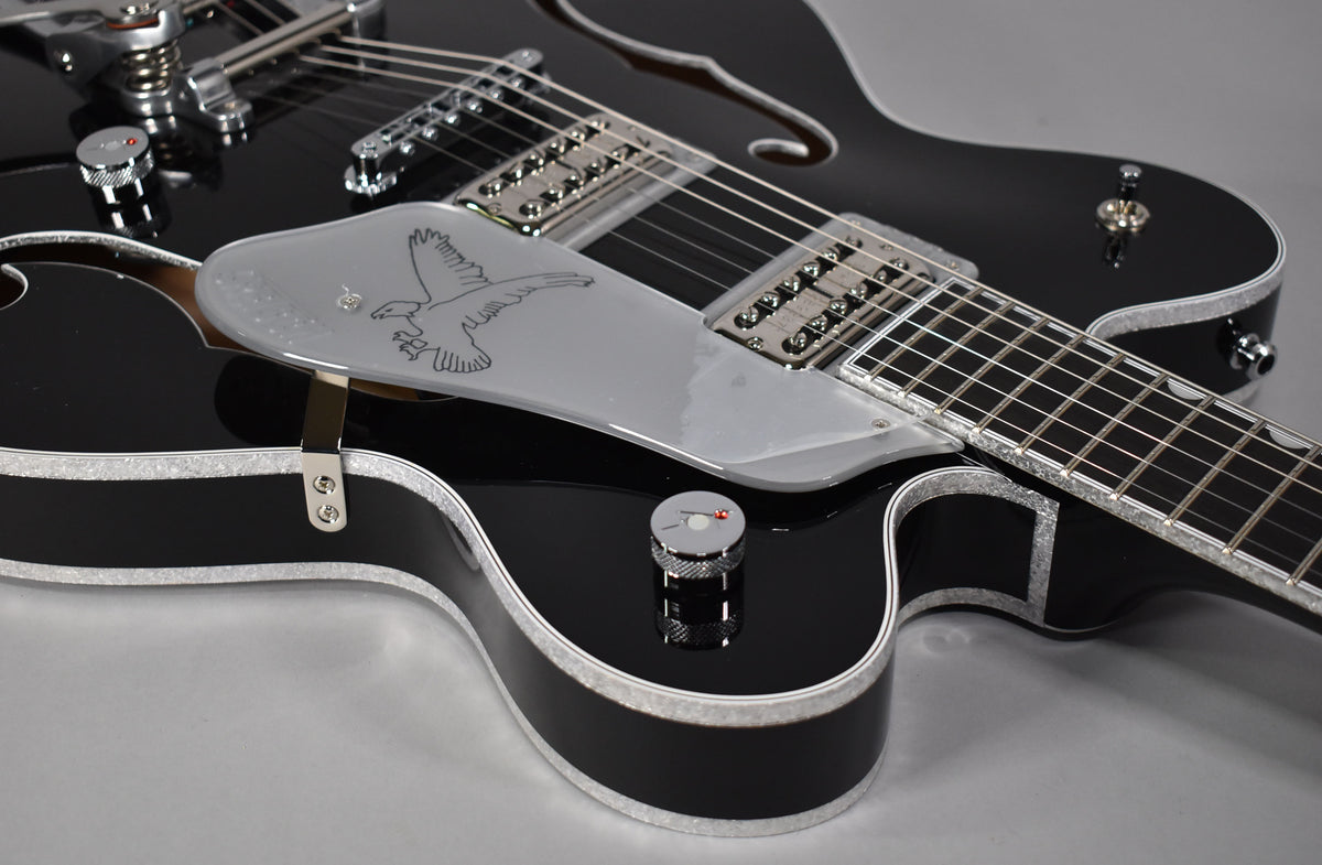 Close-up of a glossy black hollow-body electric guitar with silver sparkle binding, showing the lower bout, pickguard with eagle graphic, and chrome hardware.