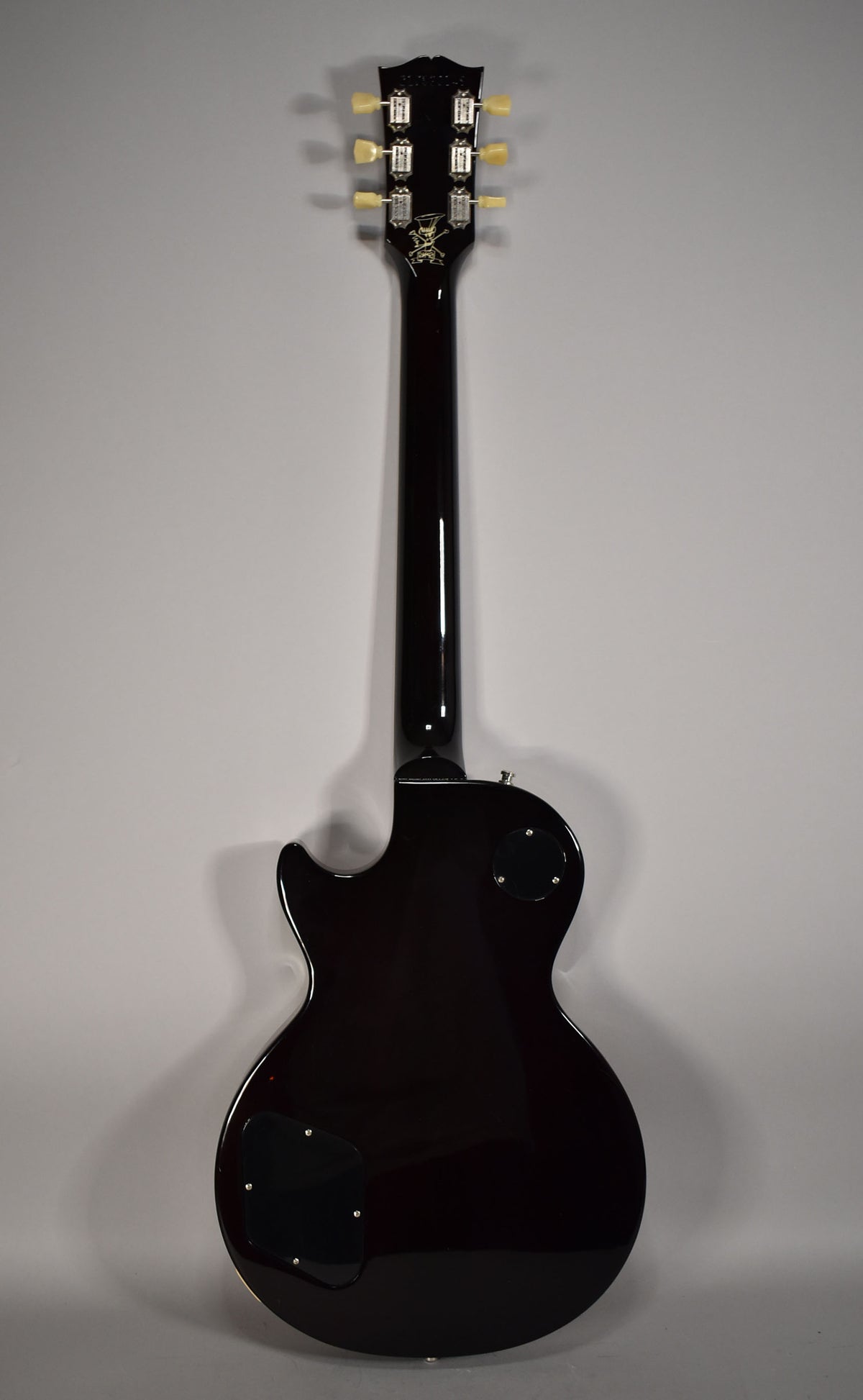 The guitar's dark brown back and neck are visible, with a black headstock, cream tuners, and a gold skull and top hat logo.