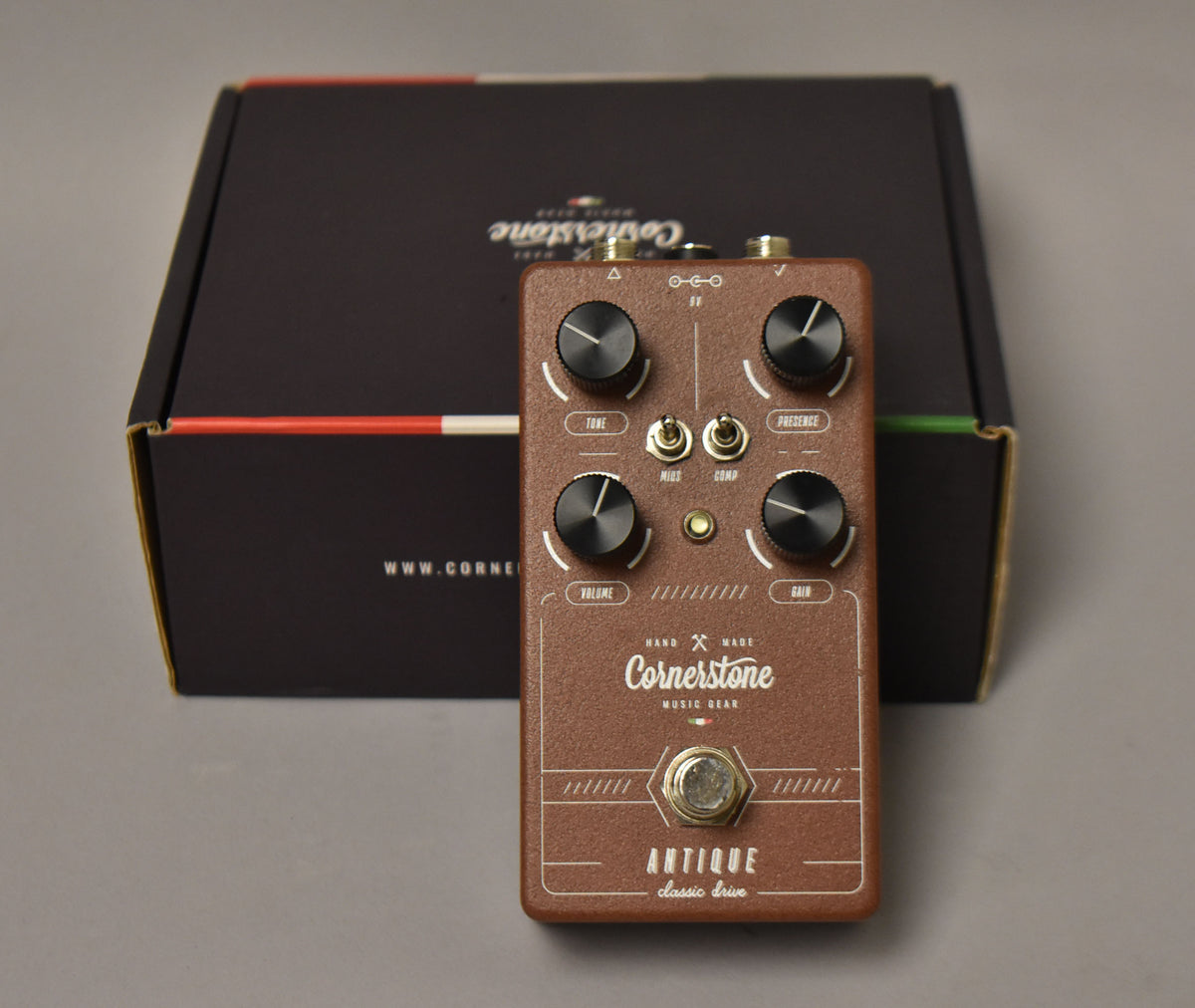 A brown guitar pedal with four black knobs, two small toggle switches, and a silver footswitch, shown with its black box.