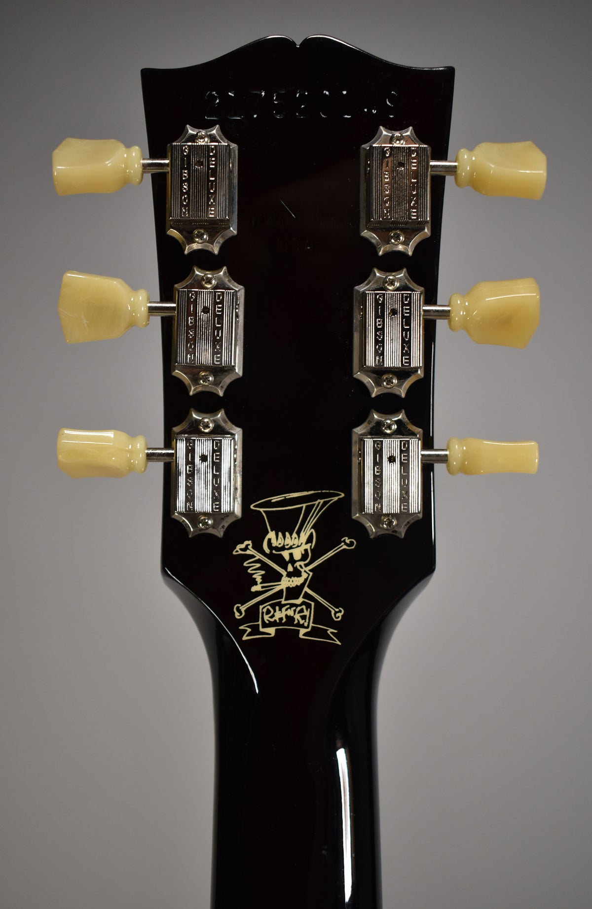 The rear of a glossy black headstock with six cream tuning pegs and a gold skull and crossbones logo.