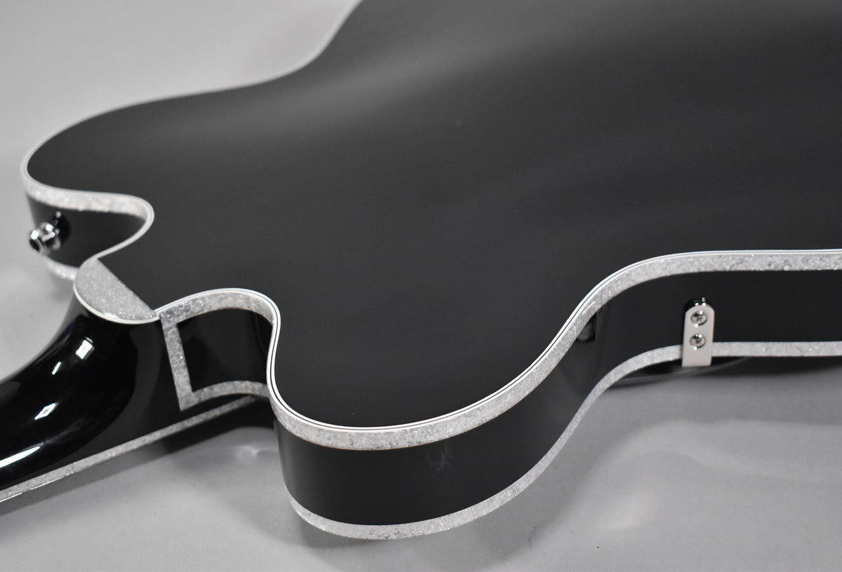 The guitar's matte black body and glossy black neck are visible from the back, with silver sparkle binding along the edges.