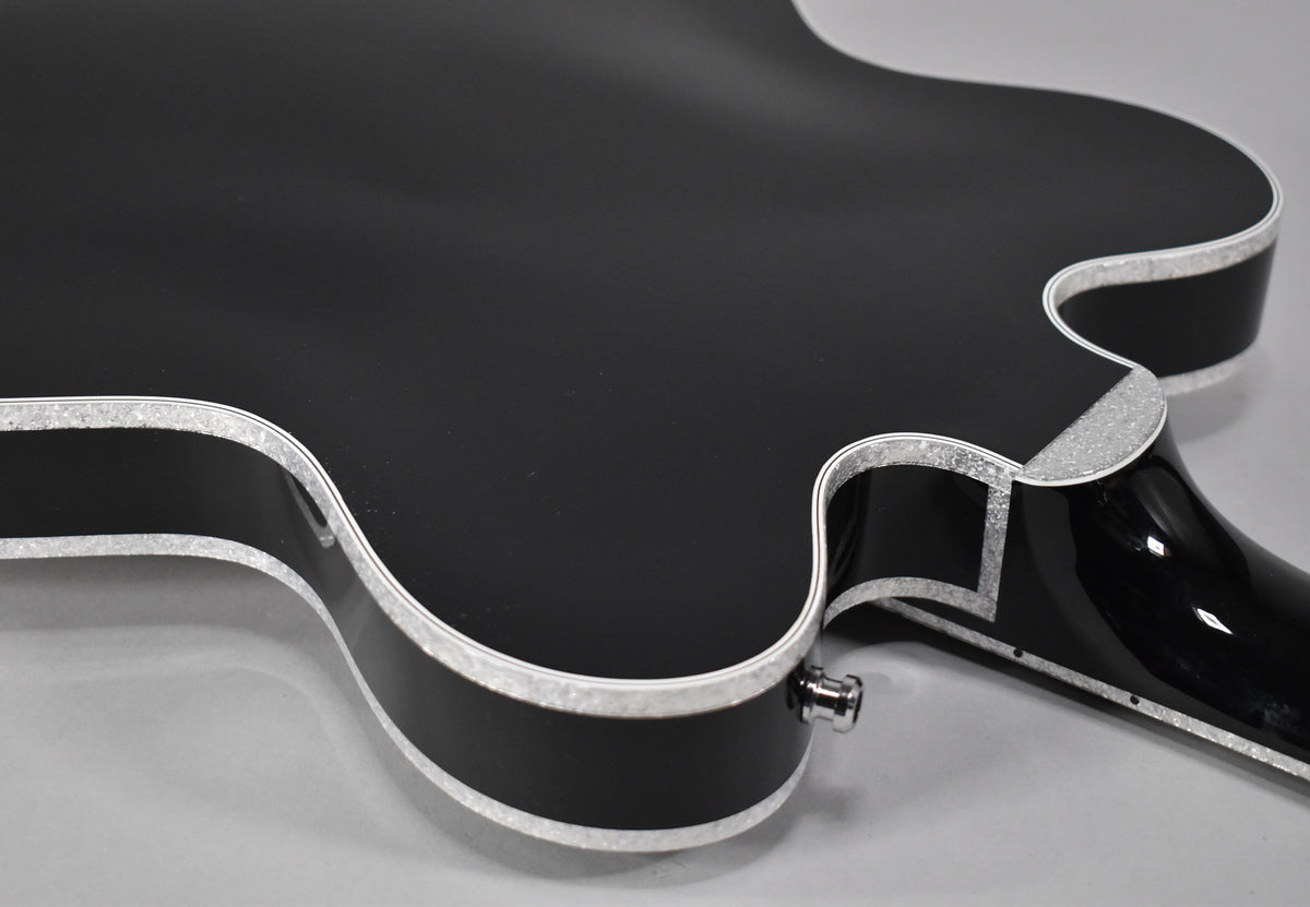 The lower bout and cutaway of a black matte hollow body guitar, with glittery silver binding and a chrome output jack.