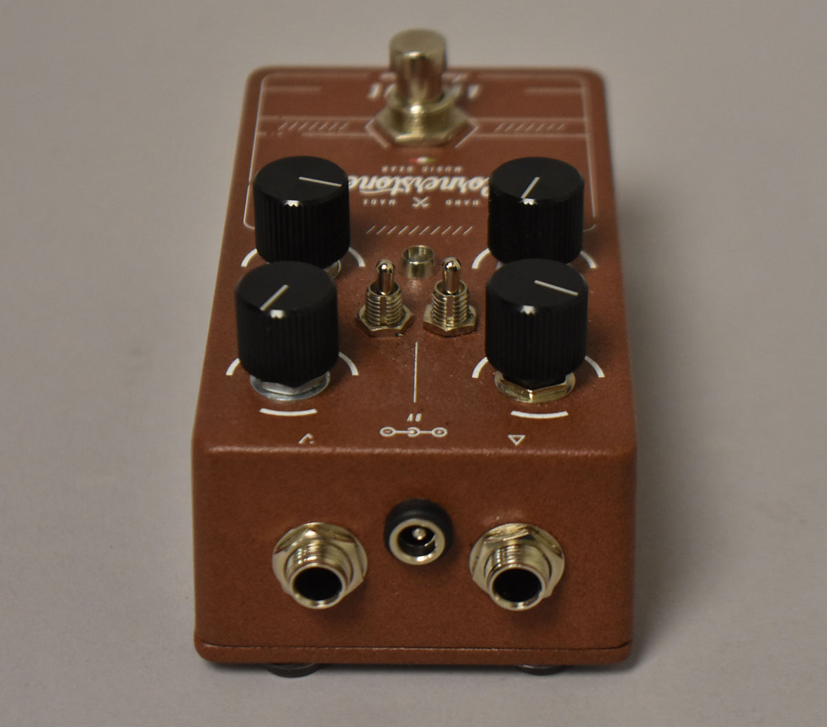 Top-down view of a brown pedal with a textured finish, four black knobs, two gold toggle switches, and three front-facing jacks.