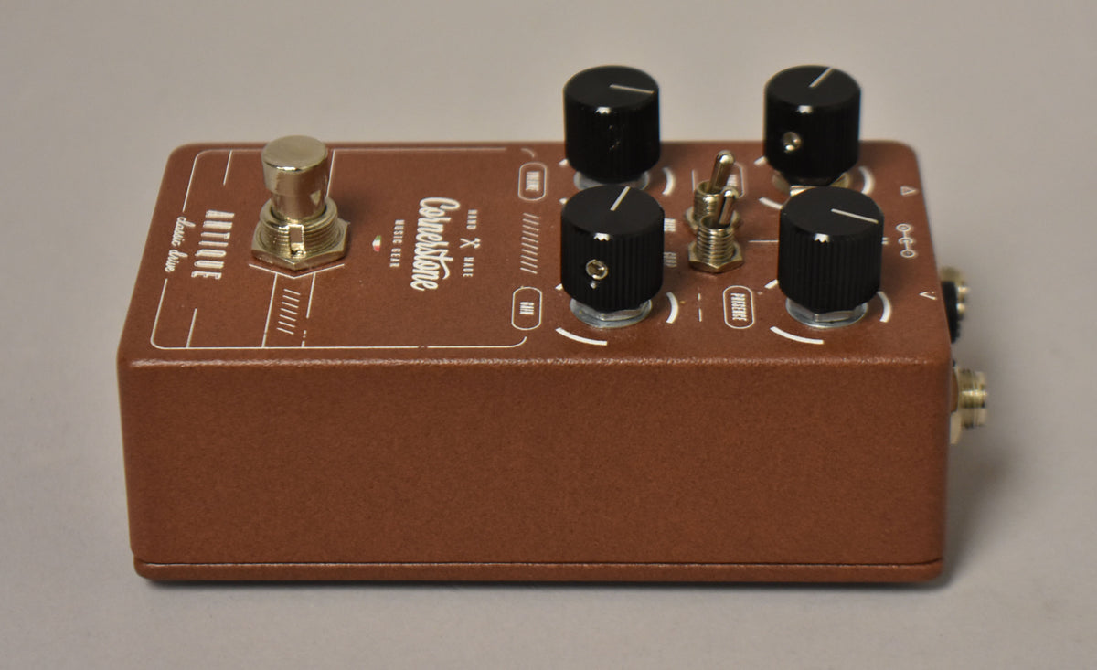 Brown pedal with a textured finish, viewed from the top right, showing a gold footswitch, four black knobs, and two mini-toggle switches.