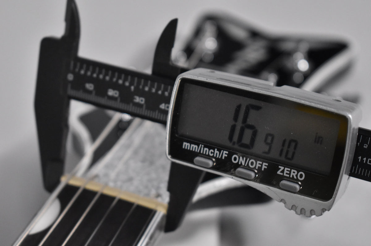 A digital caliper measures the nut width of the black guitar neck, displaying "1.6910 in" on its screen.