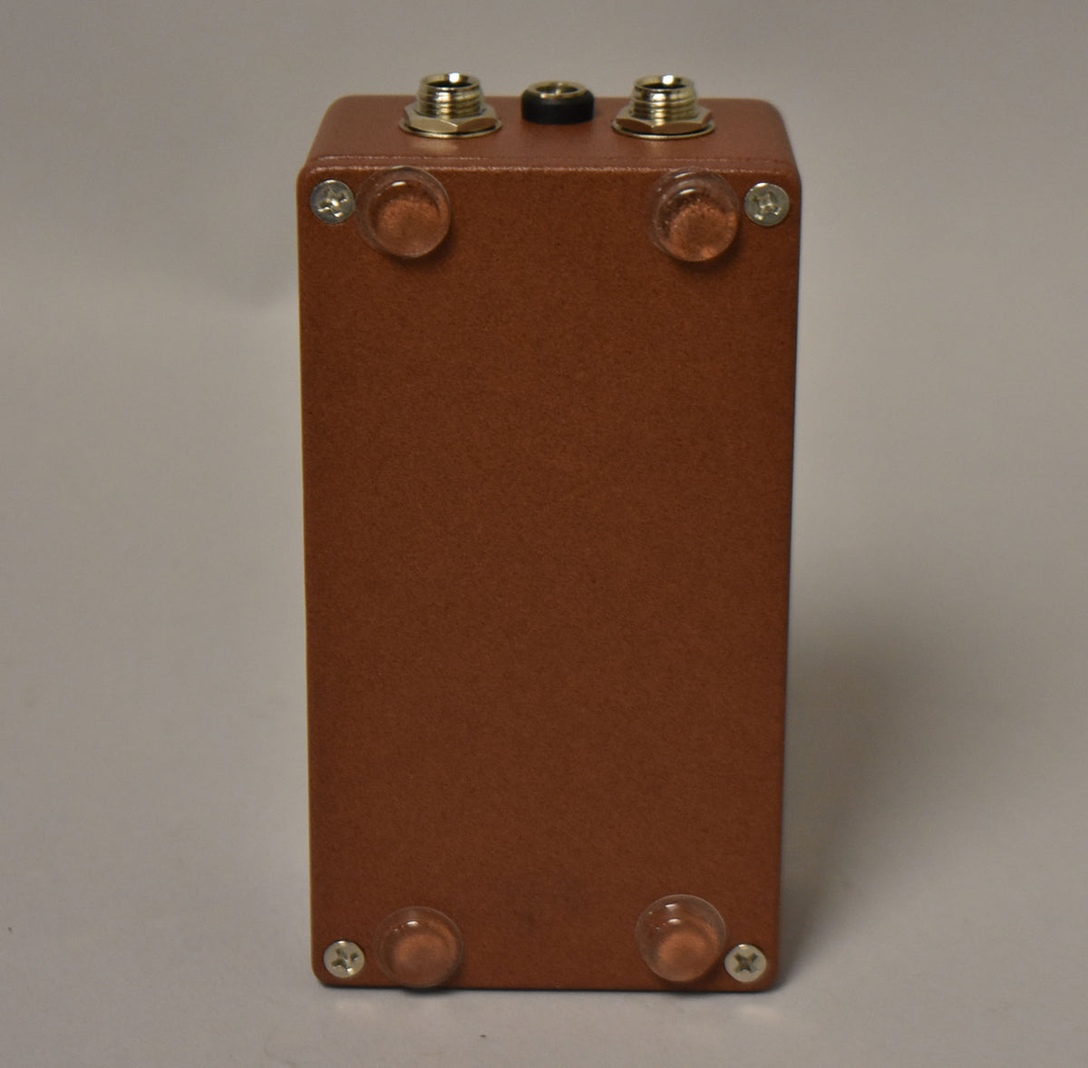 The brown pedal is viewed from the back, showing two input jacks, a power jack, and four clear rubber feet with screws.