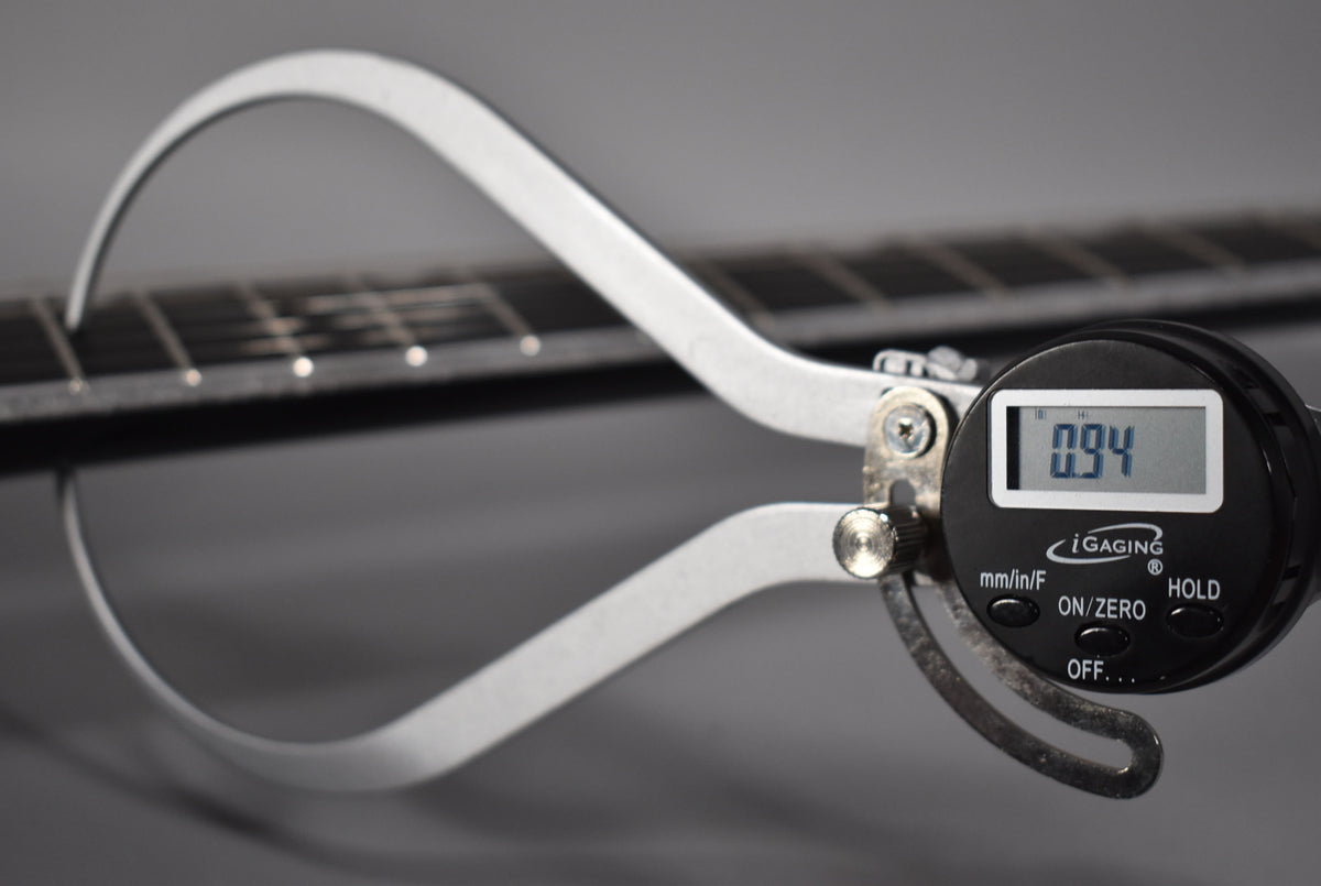 A digital caliper measures the fret height on the guitar's dark fretboard, displaying "0.94" on its screen.