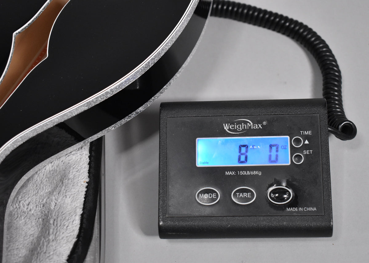 The black guitar's lower bout with silver sparkle binding rests on a scale displaying "8 lb 0 oz", with a plush case lining visible.