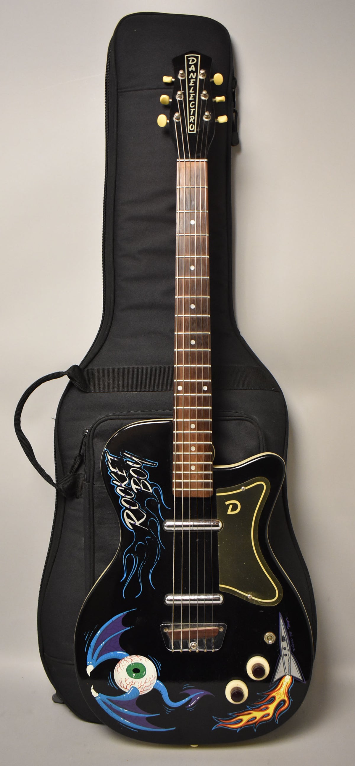 A black electric guitar with custom "Rocket Boy" art, including a winged eyeball and a rocket, rests in a black gig bag.