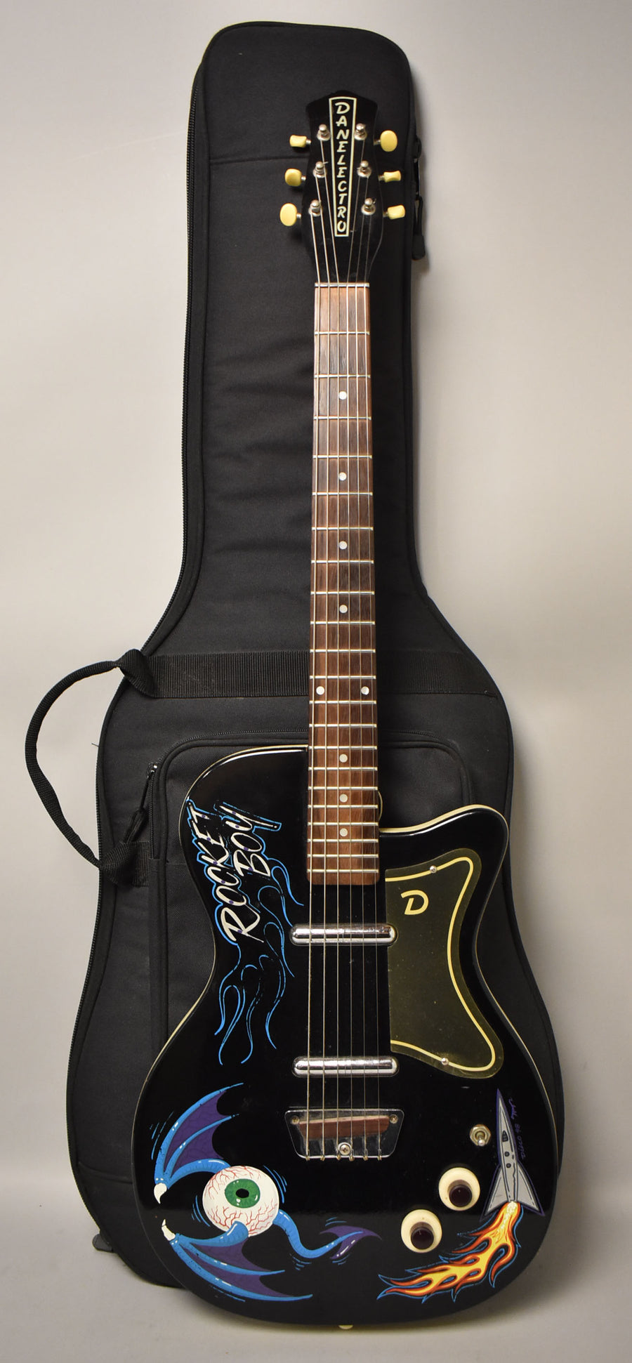 A black electric guitar with custom "Rocket Boy" art, including a winged eyeball and a rocket, rests in a black gig bag.
