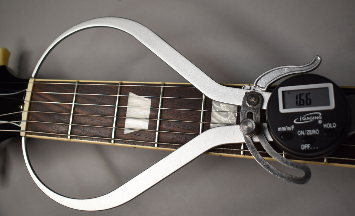 A digital caliper measures the string height at the 12th fret of the dark rosewood fretboard with trapezoid inlays.
