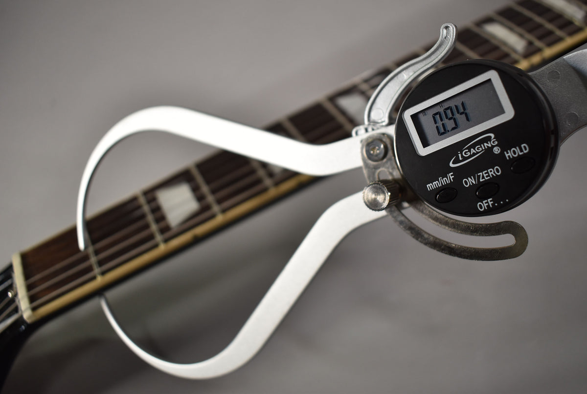 A digital caliper measures the nut width at 0.94 inches, with the guitar's dark fretboard and trapezoid inlays visible.