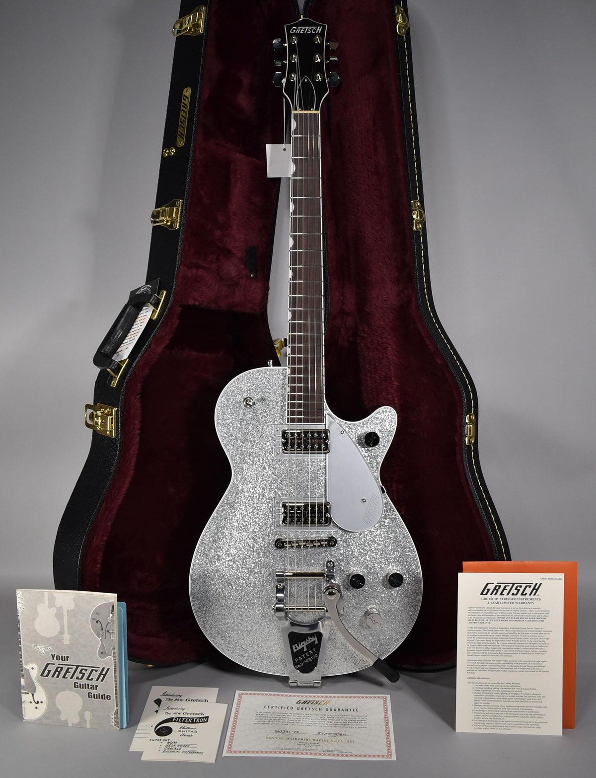 Full-length view of a silver sparkle electric guitar with a Bigsby tremolo, two humbuckers, and a silver pickguard, standing in its open, red-lined hardshell case.