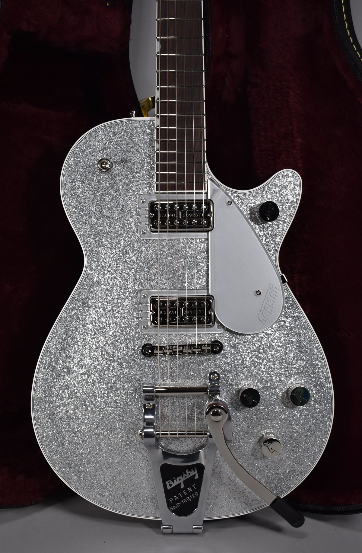 The silver sparkle guitar is angled slightly, showing its Bigsby vibrato, two humbuckers, silver pickguard, and four black control knobs.
