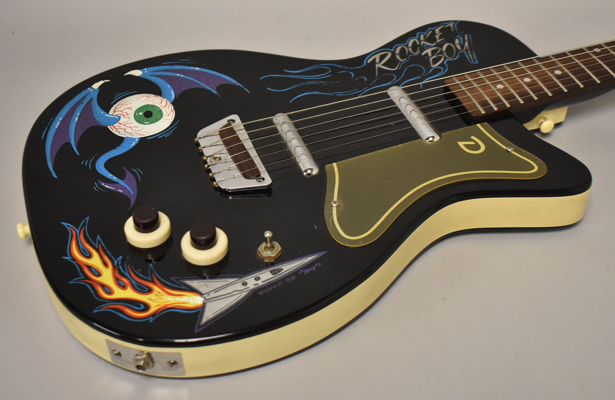 Black electric guitar, angled from the upper right, showing custom painted flames, a winged eyeball, and a rocket ship.