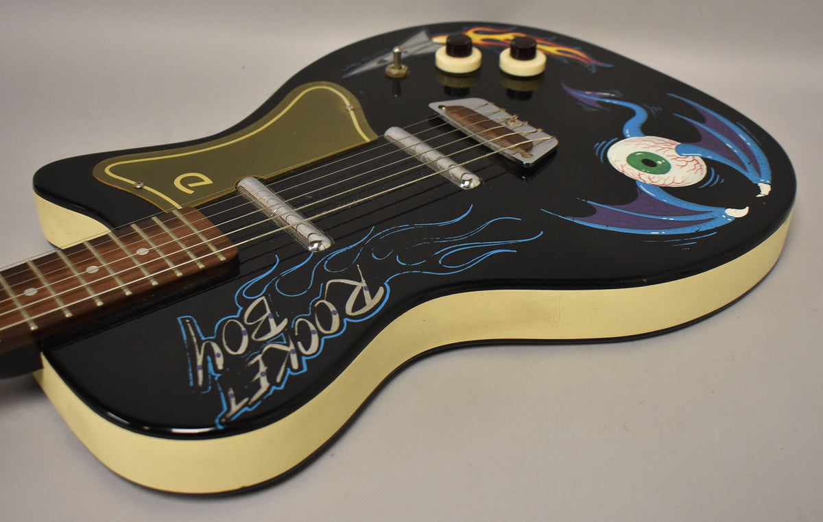 The black guitar body, with cream sides, features custom "Rocket Boy" lettering, blue flames, and a winged eyeball graphic.