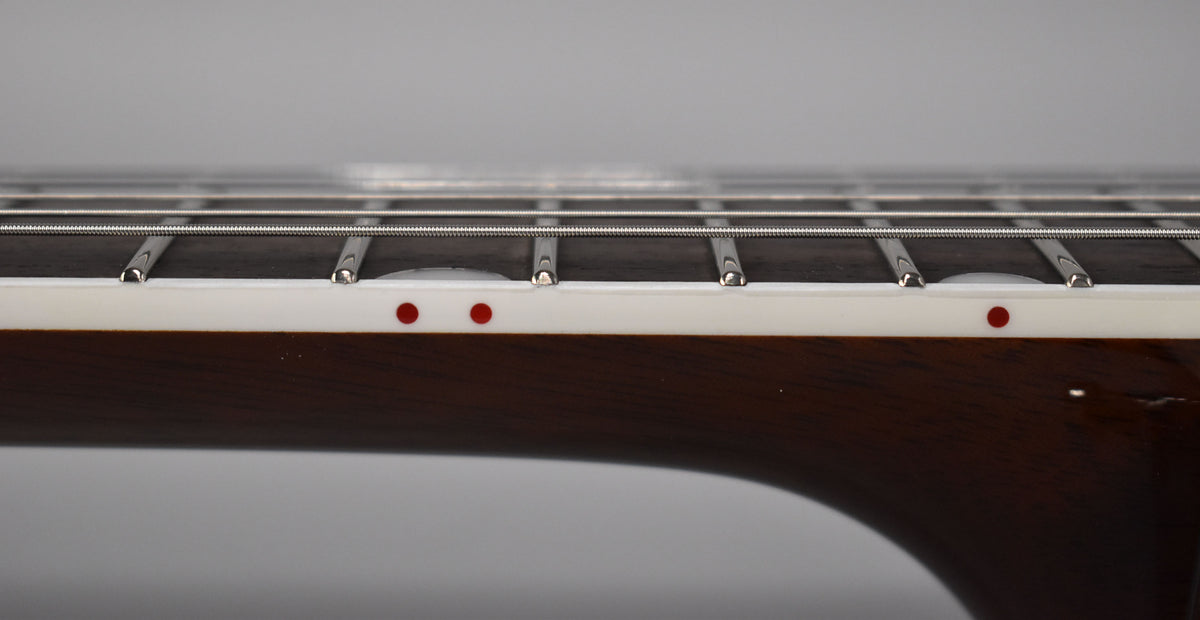 The guitar's dark fretboard, with silver frets and strings, is framed from the side, showing white binding with red dot markers.