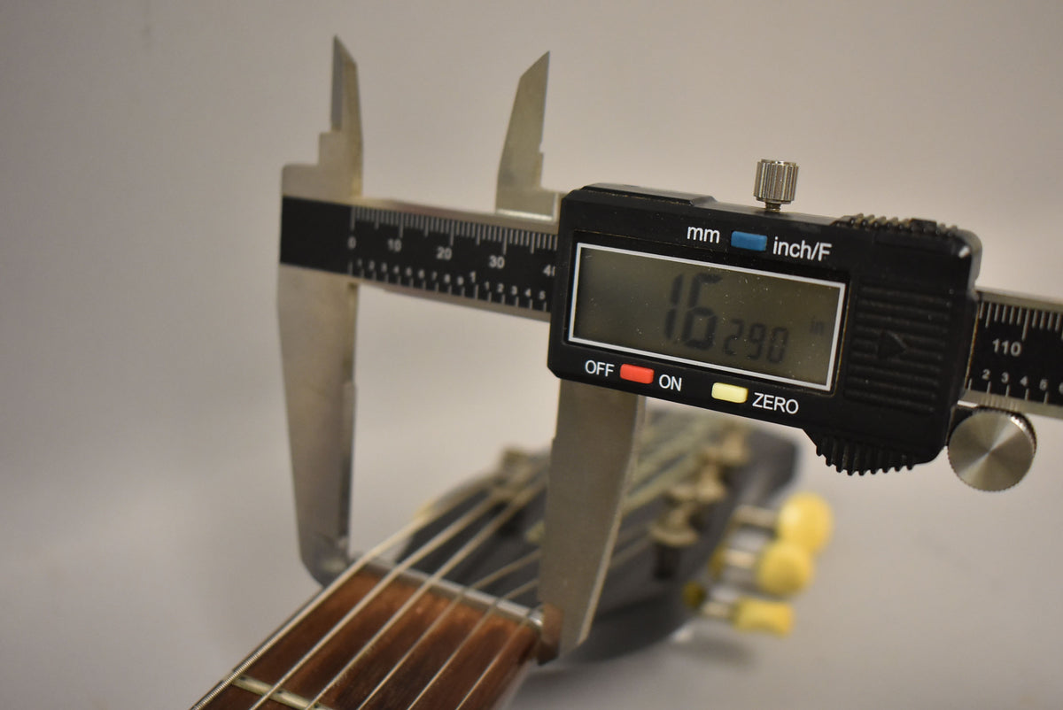 A digital caliper measures the string height at the first fret, showing 1.629mm, with the headstock and tuners blurred in the background.