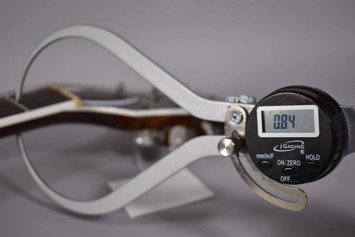 A digital caliper measures the guitar's neck, displaying "0.84" on its screen, with the fretboard visible in the background.