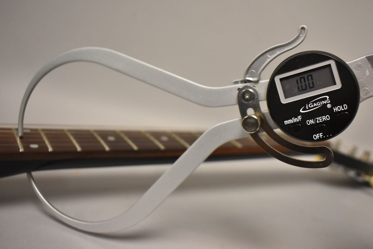 A digital caliper measures the string height at the first fret of a dark fretboard with light fret markers.