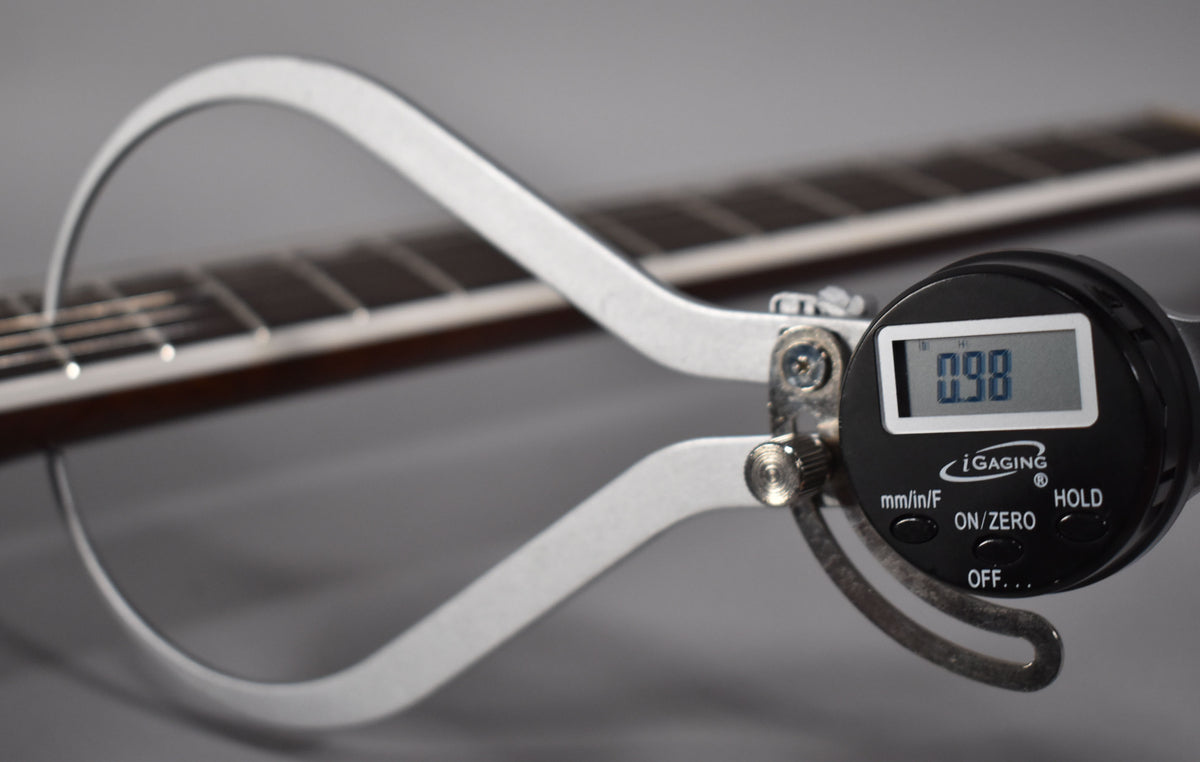 A digital caliper measures the fret height on the guitar's dark fretboard, displaying "0.98" on its screen.