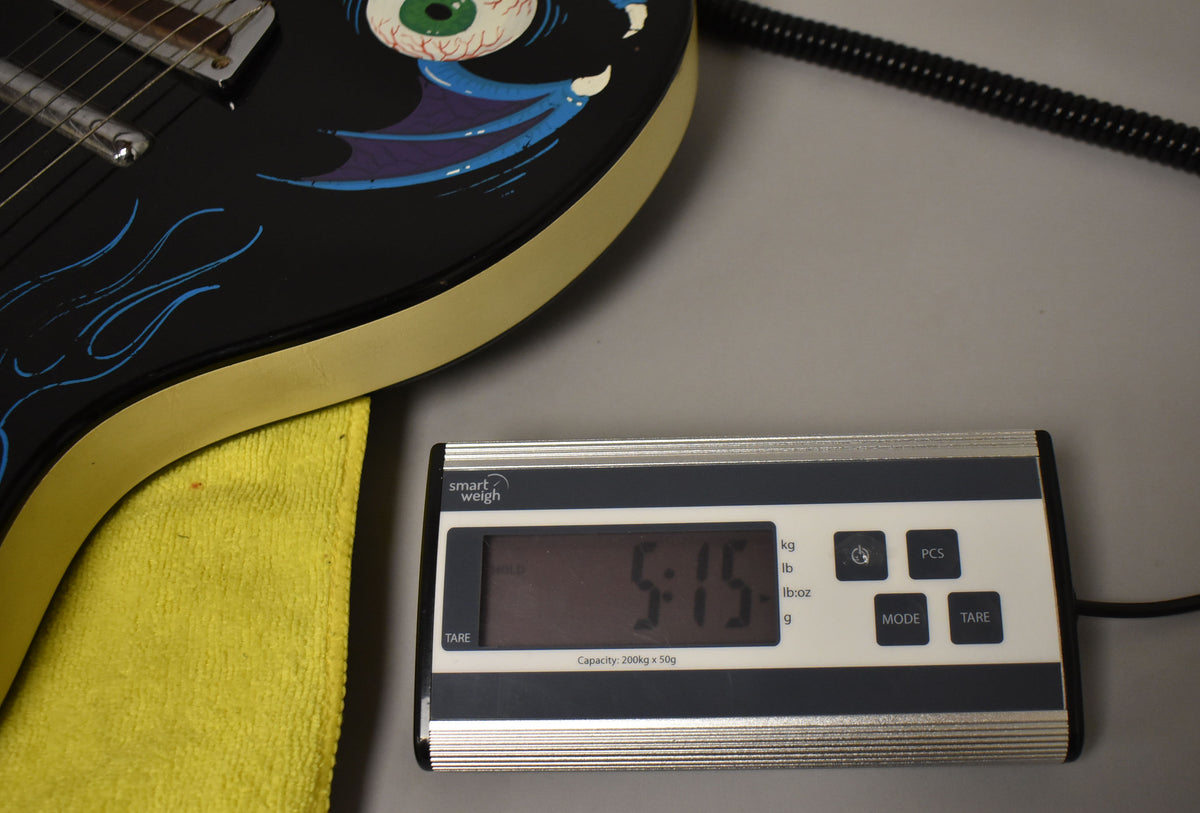 The guitar's black body with custom blue flames and a green eyeball graphic rests on a scale displaying "5.15" lbs.