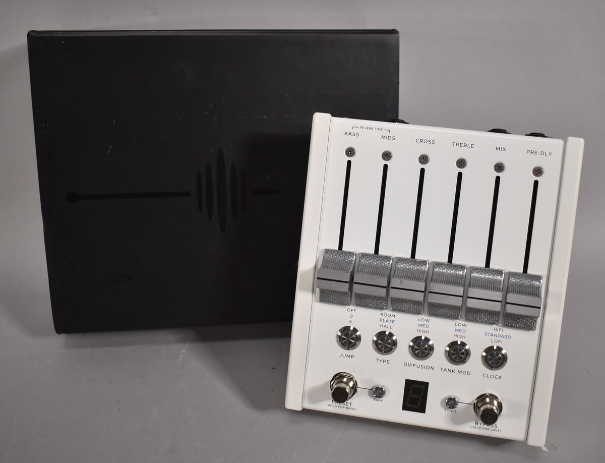 White effect pedal with six silver faders, six rotary knobs, two footswitches, and a small digital display, next to a black box.