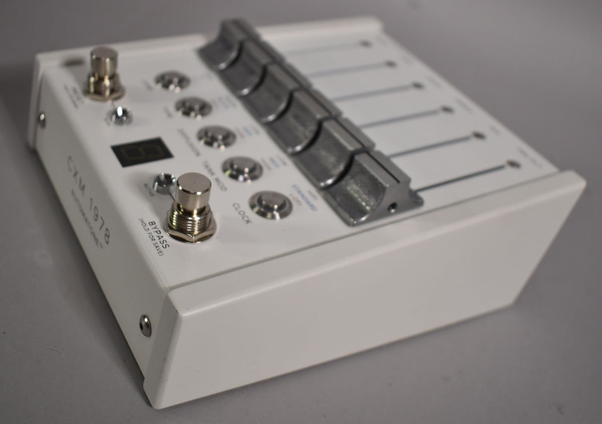 A white effect pedal is angled from the right, showing its top with two footswitches, a digital display, and five metal faders.