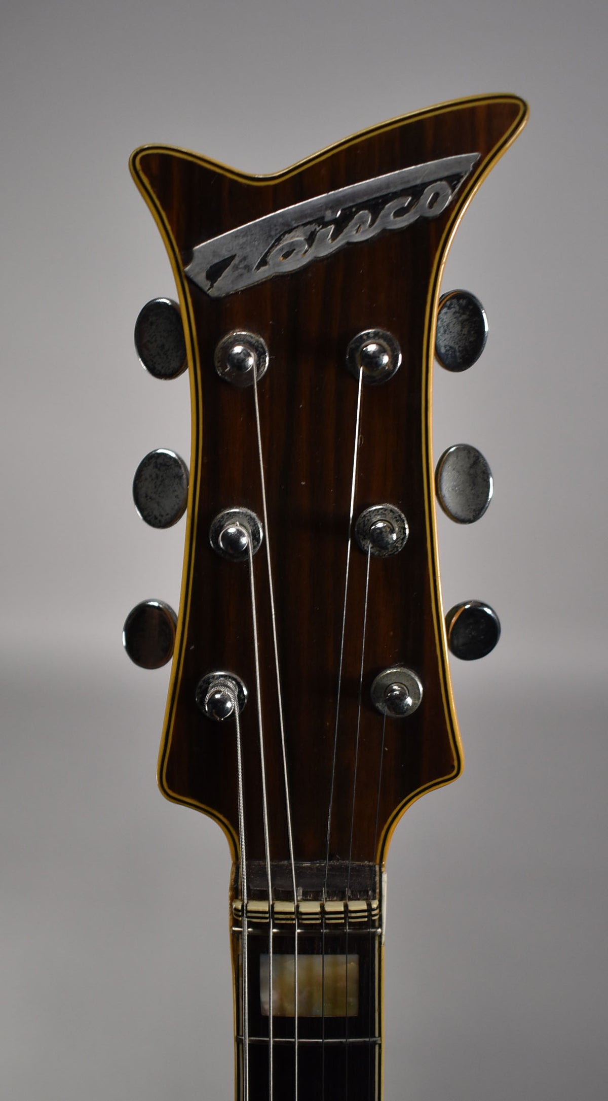 Close-up of the dark wood headstock with a silver logo, six tarnished tuners, and a portion of the fretboard with block inlays.