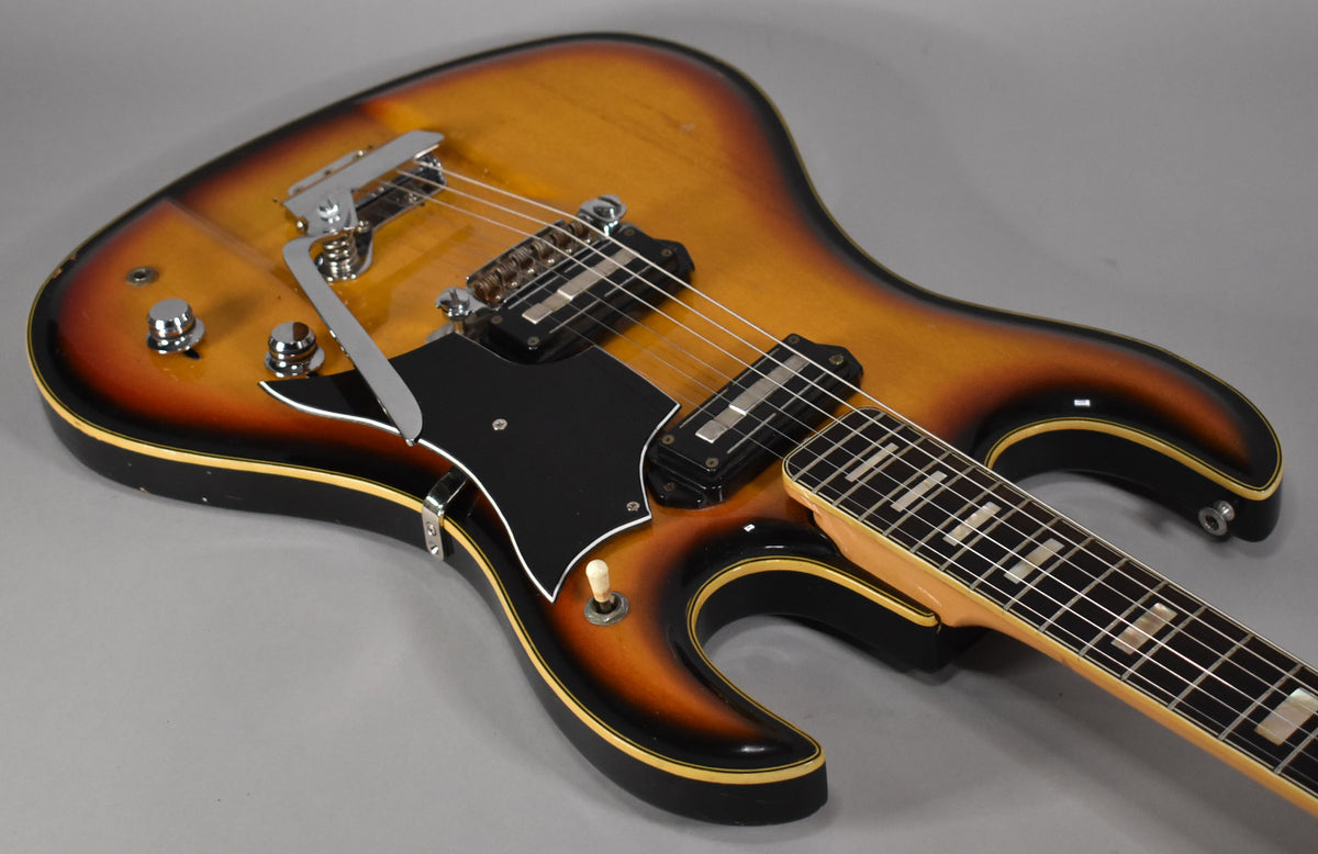 Angled view of a sunburst guitar body with a vibrato tailpiece, two pickups, black pickguard, and a three-way switch.