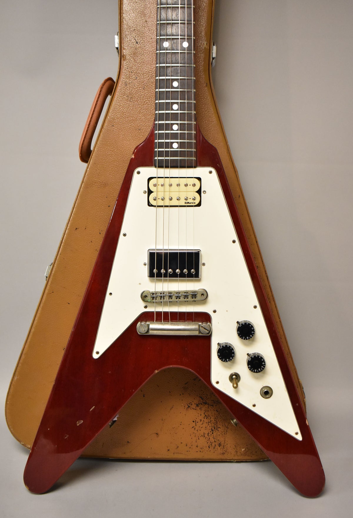 A cherry red Flying V electric guitar with a white pickguard, two humbuckers, and three black control knobs, resting in its brown case.