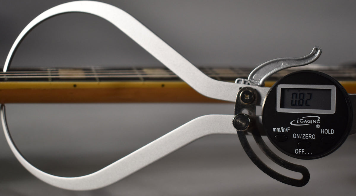 A digital caliper measures the guitar's neck, showing a reading of 0.82, with the fretboard and strings visible.
