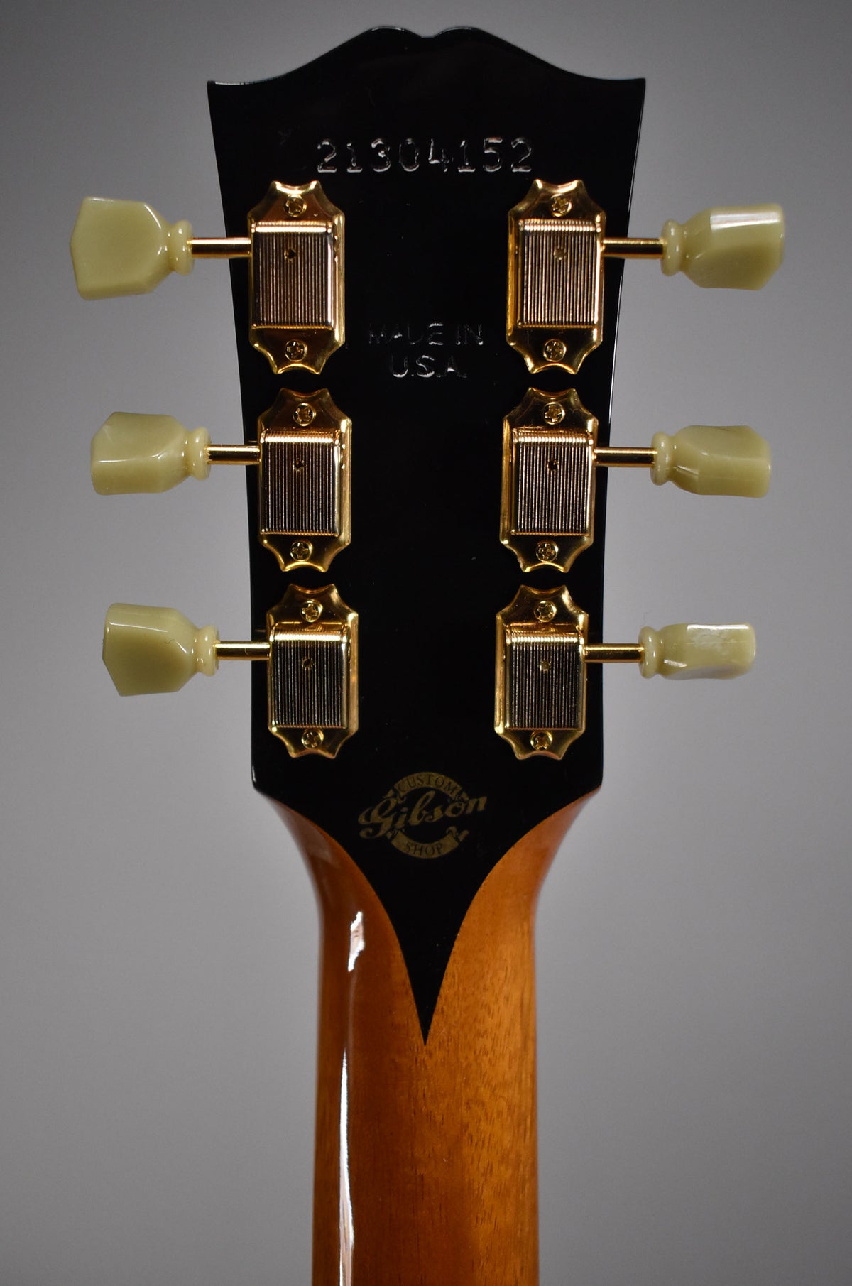 The back of the guitar's headstock, with a glossy black finish, six gold tuners with cream buttons, and a natural wood neck.
