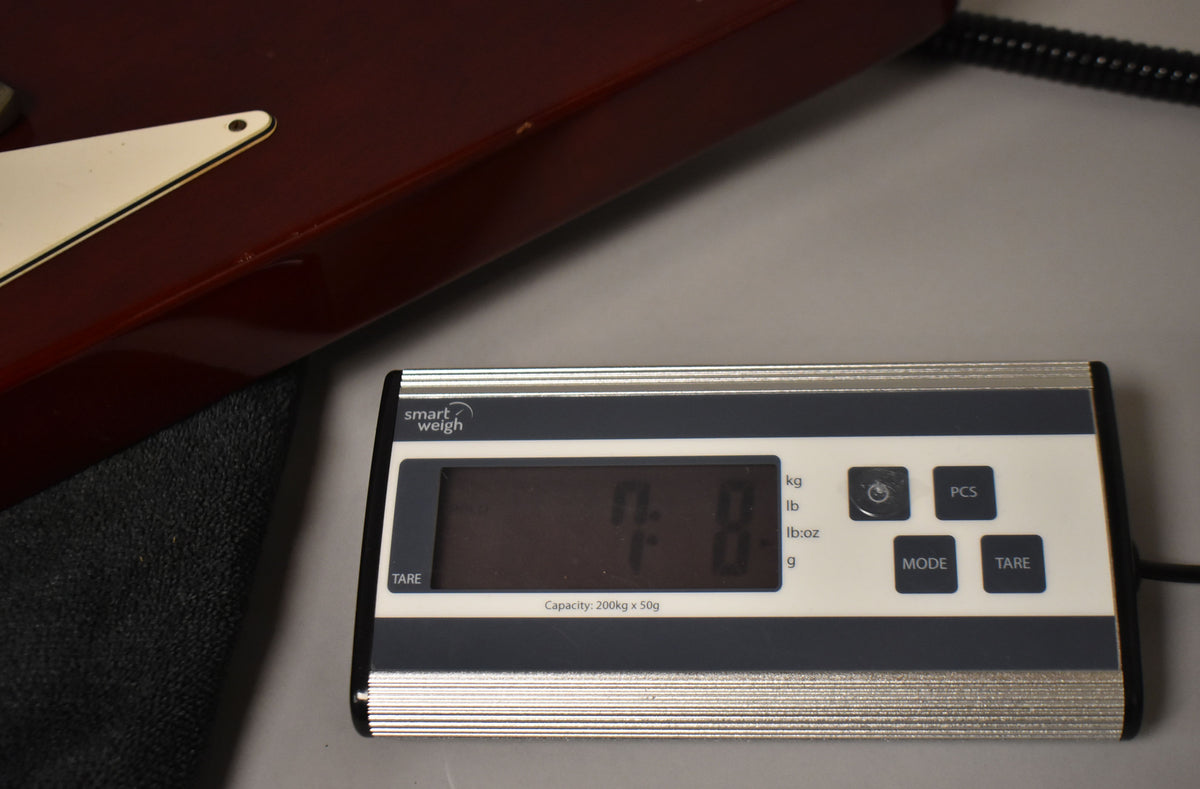 The cherry red Flying V body, with a cream pickguard, rests on a scale displaying "7.8" pounds.