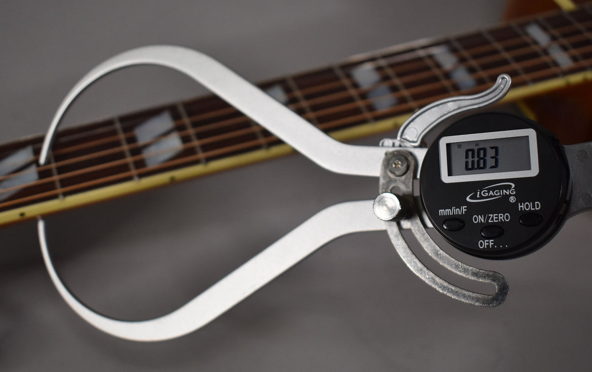 A digital caliper measures the string height at the 12th fret of the guitar's rosewood fretboard with trapezoid inlays.