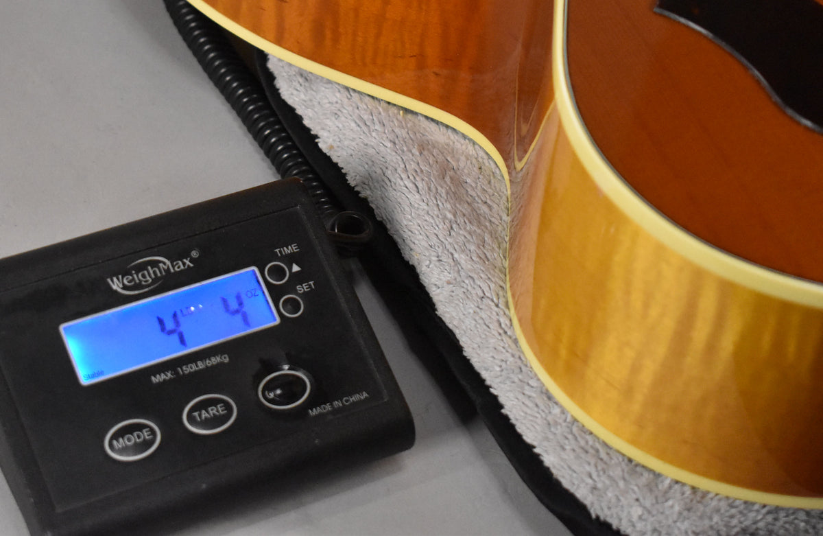 The guitar's lower bout and waist are shown on a scale displaying "4 lbs 4 oz", with a plush gray case lining.