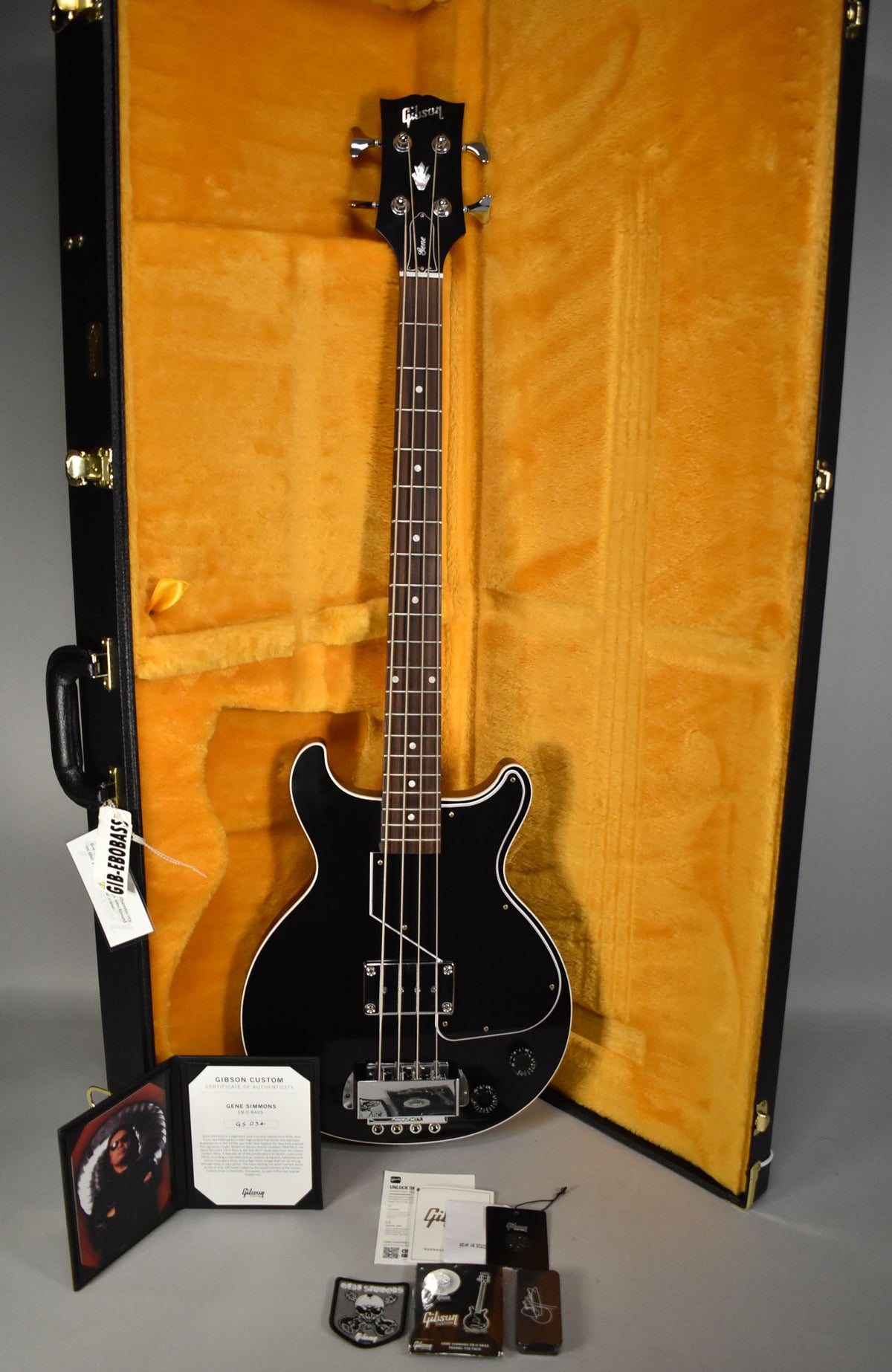 The black bass with white binding and chrome hardware lies in its open, gold-lined hard case, with paperwork and accessories in front.
