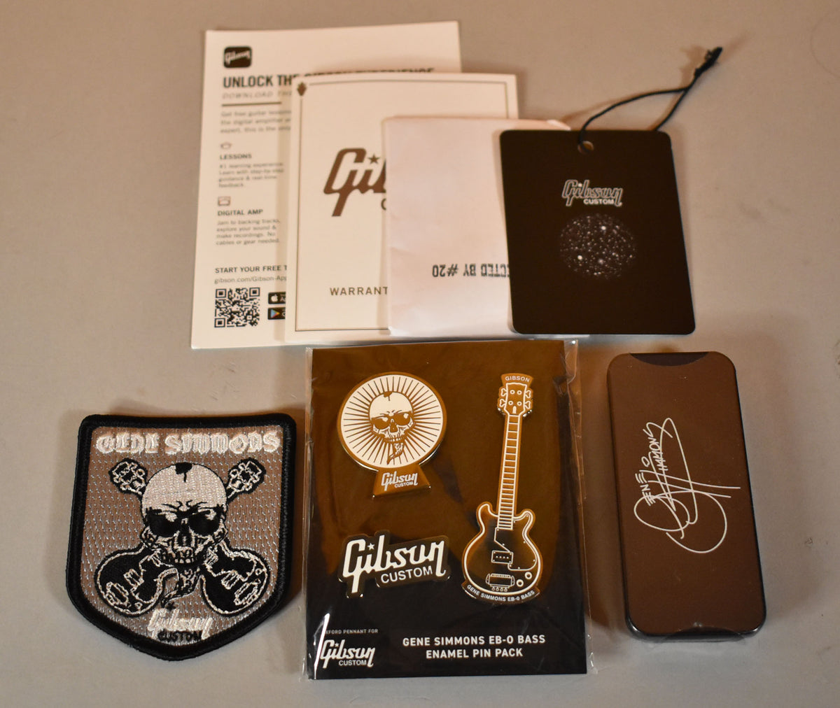 Assorted Gene Simmons-themed accessories are shown, including a patch, enamel pin pack, and a signed backplate.