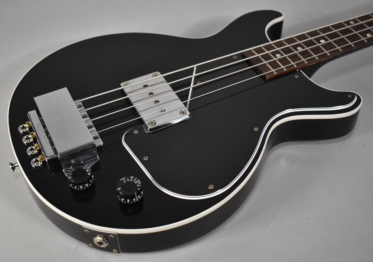 The black bass guitar, viewed from the upper right, has a chrome bridge and pickup, black pickguard, and white binding along the body.