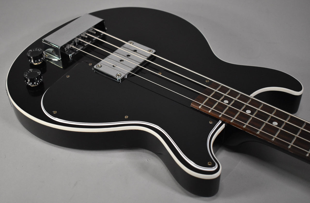 The black bass guitar, with white binding and a chrome bridge cover, is angled to show the body, controls, and fretboard.