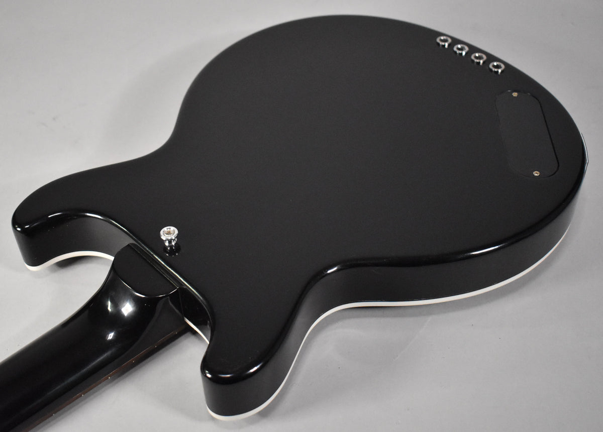 The bass's glossy black body, with white binding, is seen from the back, angled to show the neck joint and control cavity.