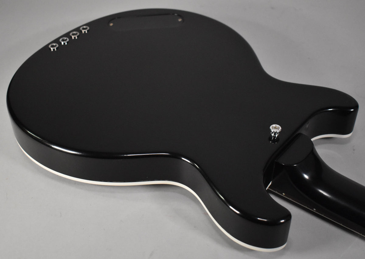 The bass's glossy black body, with white binding, is seen from the back, angled to show the neck joint and four chrome string ferrules.