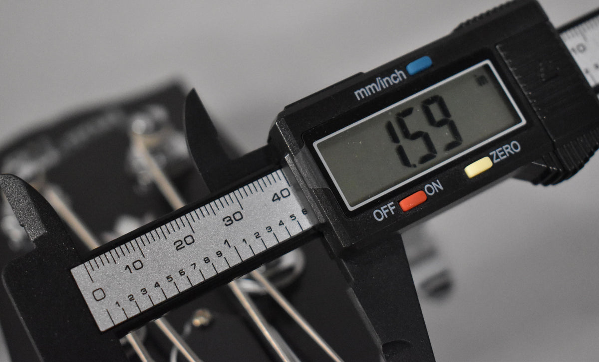 A digital caliper measures the bass's nut width at 1.59 inches, with the headstock and strings visible in the background.