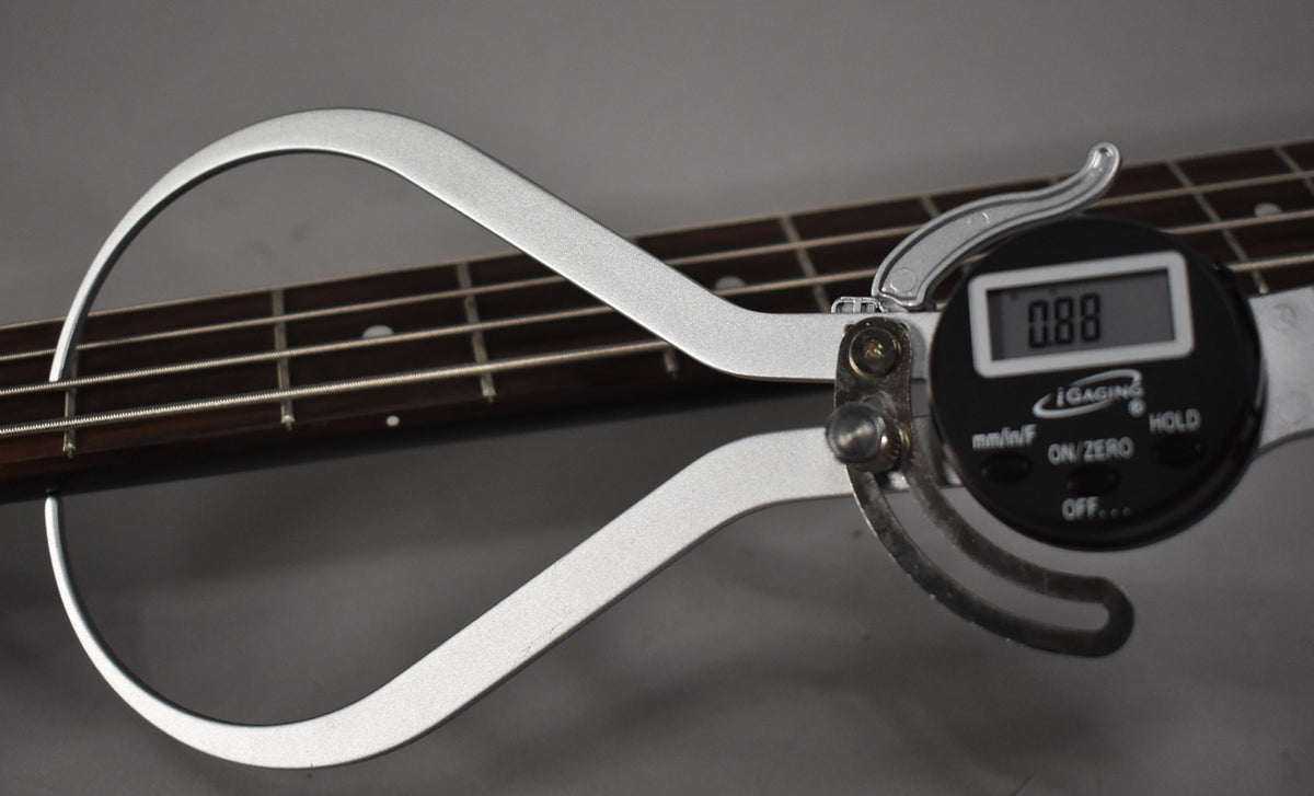 A digital caliper measures the bass's dark fretboard and strings, displaying "0.88" on its black screen.