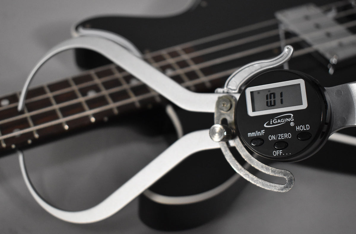 A digital caliper measures the string height at 1.01, resting on the ebony bass's dark fretboard and black body.