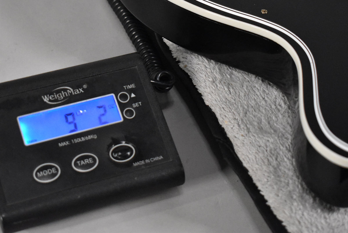 The bass rests in its case, with a digital scale displaying "9.2" lbs next to its black body with white binding.